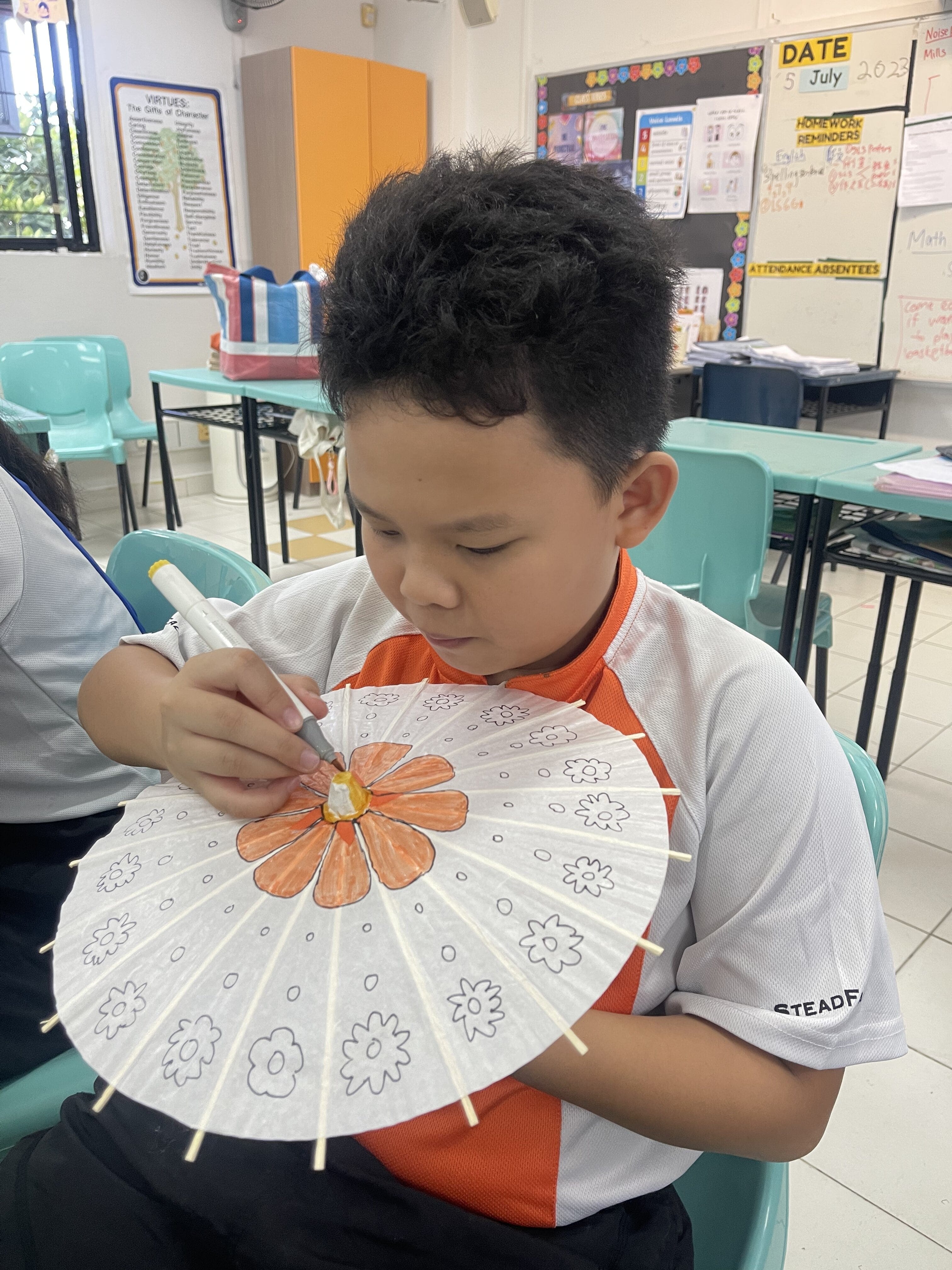 A young boy in a classroom colors an orange flower on a small white paper parasol with a marker.